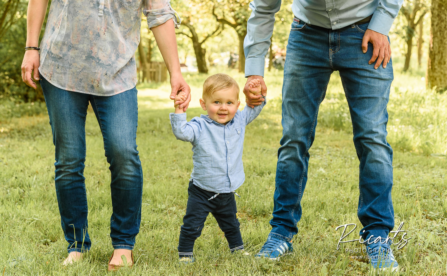 junge-Familie-mit-Kleinkind-an-den-Händen-geht-auf-Wiese-spazieren