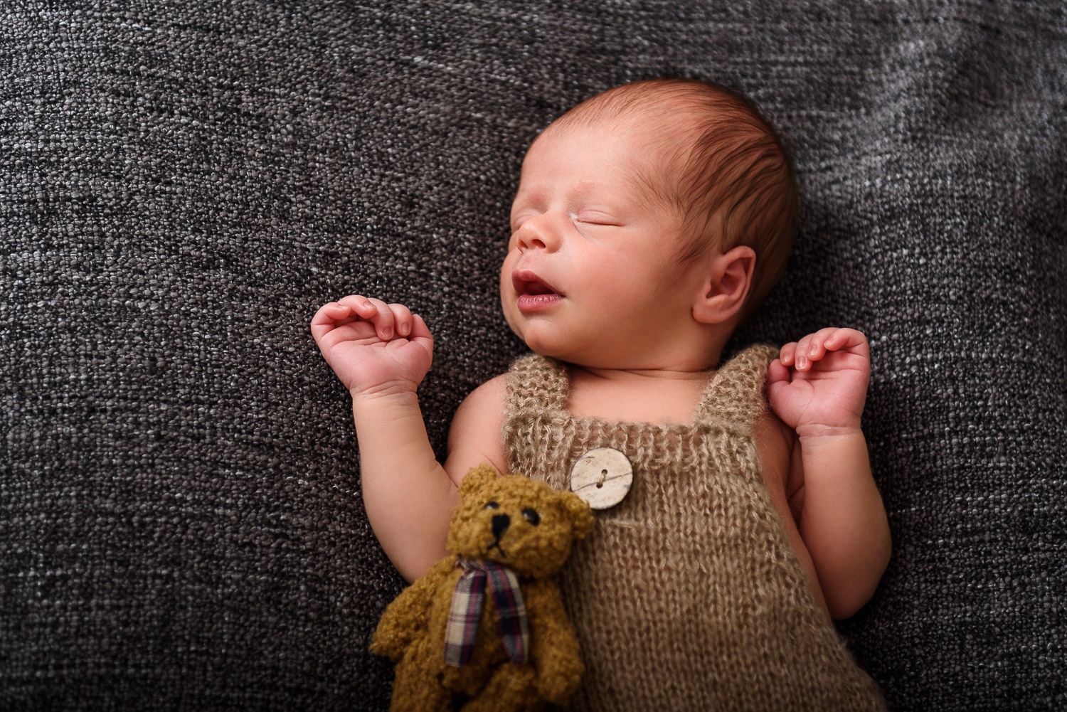 Neugeborenes-Baby-liegt-schlafend-auf-einer-dunklen-Decke-mit-kleinem-Teddybaer