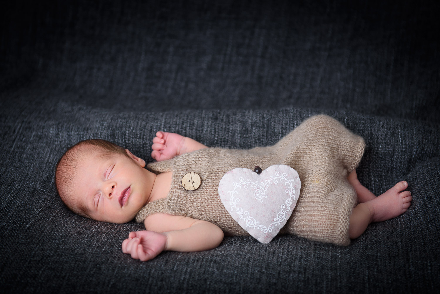 Neugeborenes-Baby-liegt-schlafend-auf-einer-dunklen-Decke-mit-Stoffherz-als-Deko