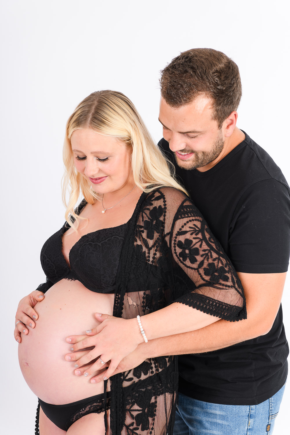 Junger-Mann-umarmt-seine-schwangere-Frau-von-Hinten-und-haelt-Haende-auf-den-Bauch