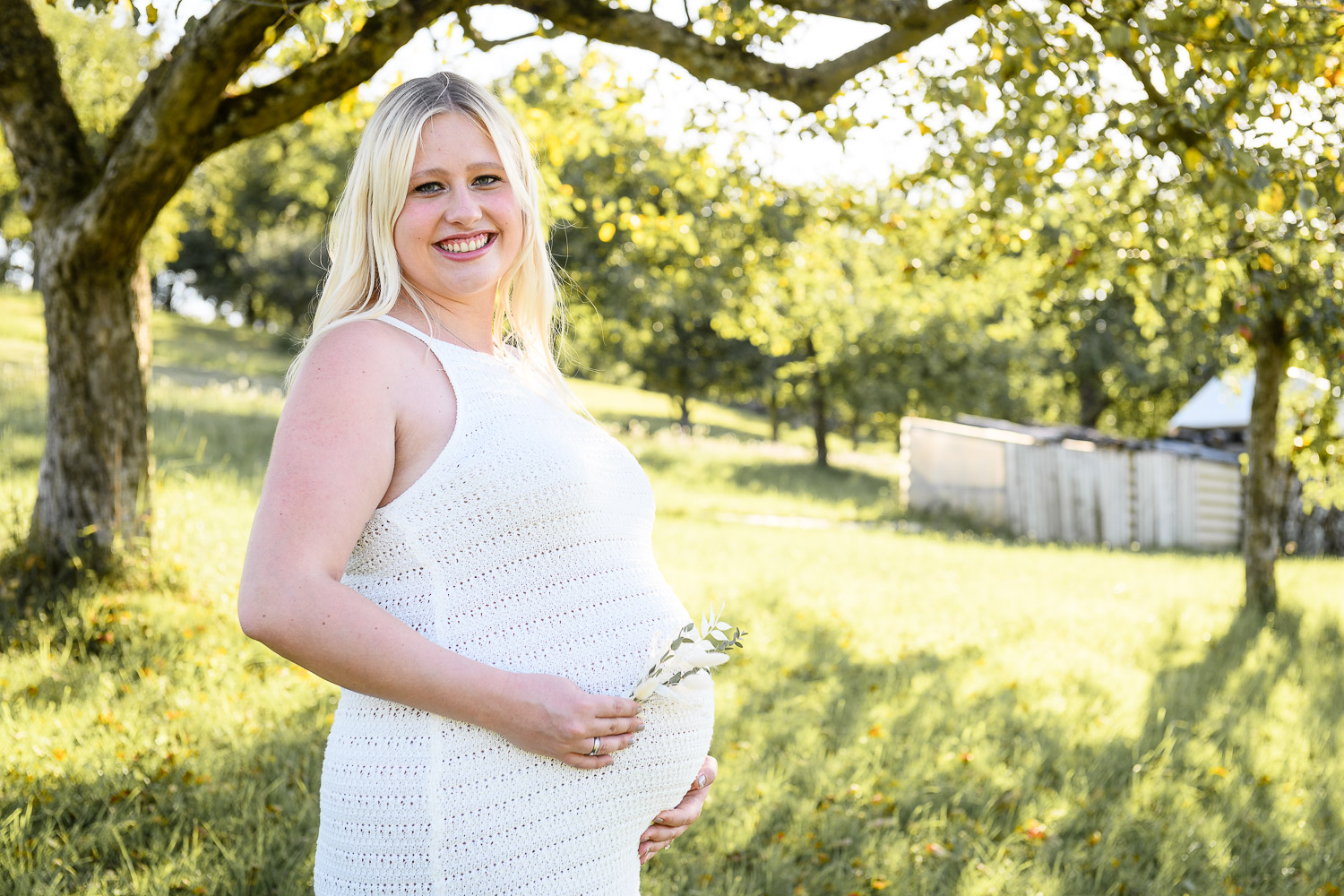 Junge-schwangere-Frau-steht-im-Sommerleid-auf-einer-Wiese-mit-Baeumen-und-lacht-in-Kamera
