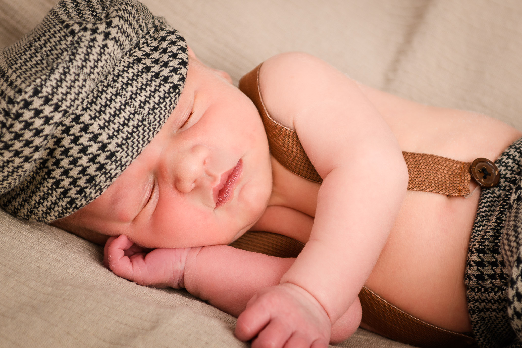 Neugeborenes-Baby-mit-Schildkappe-und-Hosenträgerhose-liegt-schlafend-auf-einem-hellen-Hintergrundstoff